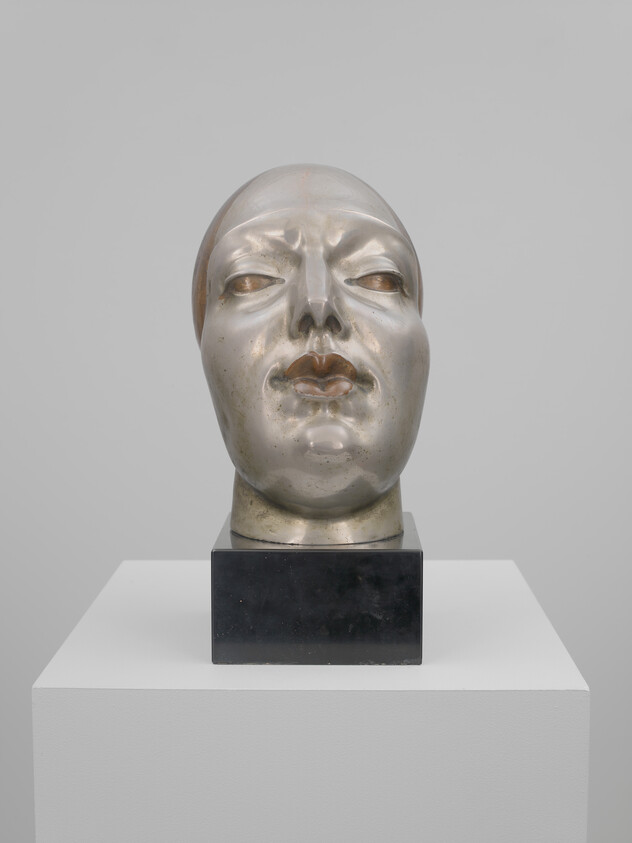 A shiny metallic human head sculpture with closed eyes displayed on a black pedestal.