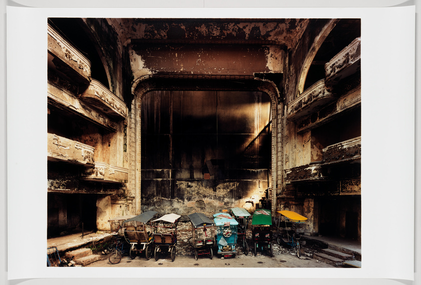 A photograph of the interior of a dilapidated theater with peeling paint and visible decay. The grand proscenium arch frames the dark, empty stage area. In the foreground, several colorful rickshaws are parked haphazardly, contrasting with the faded grandeur of the theater's architecture.