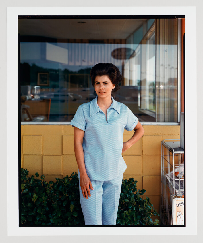 Woman in blue patterned uniform standing by a yellow wall outside a diner, with a reflection in the window and a birdcage nearby.