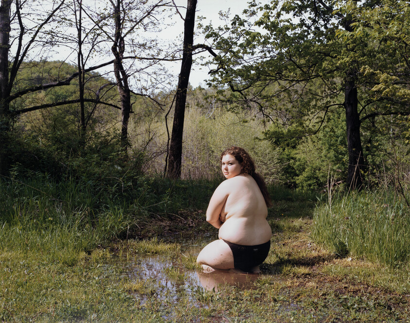 A person wearing black shorts kneels in a shallow puddle in a grassy forest clearing.