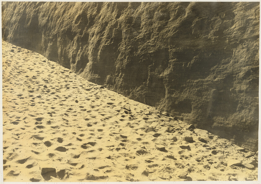 Sandy beach covered in many footprints next to a tall, textured rock cliff.