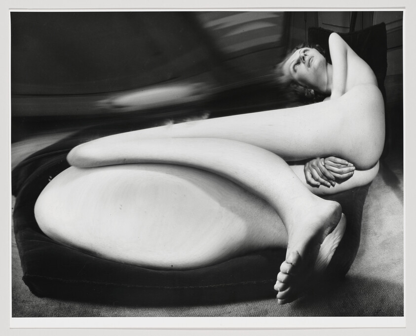 Nude person curled on a cushion with legs crossed and hands clasped near hips.