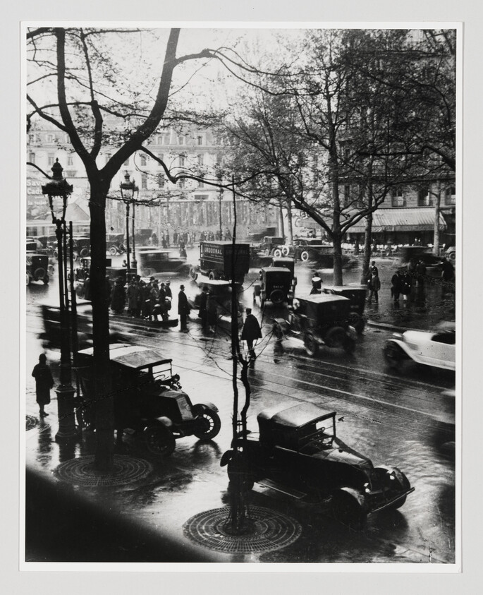 Vintage cars and pedestrians navigate a wet city street under bare trees and streetlamps.