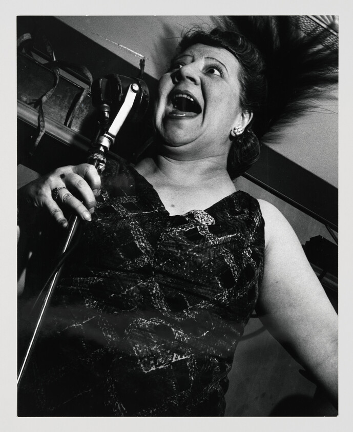 A woman in a patterned dress sings passionately into an old-fashioned microphone with a raised hand.