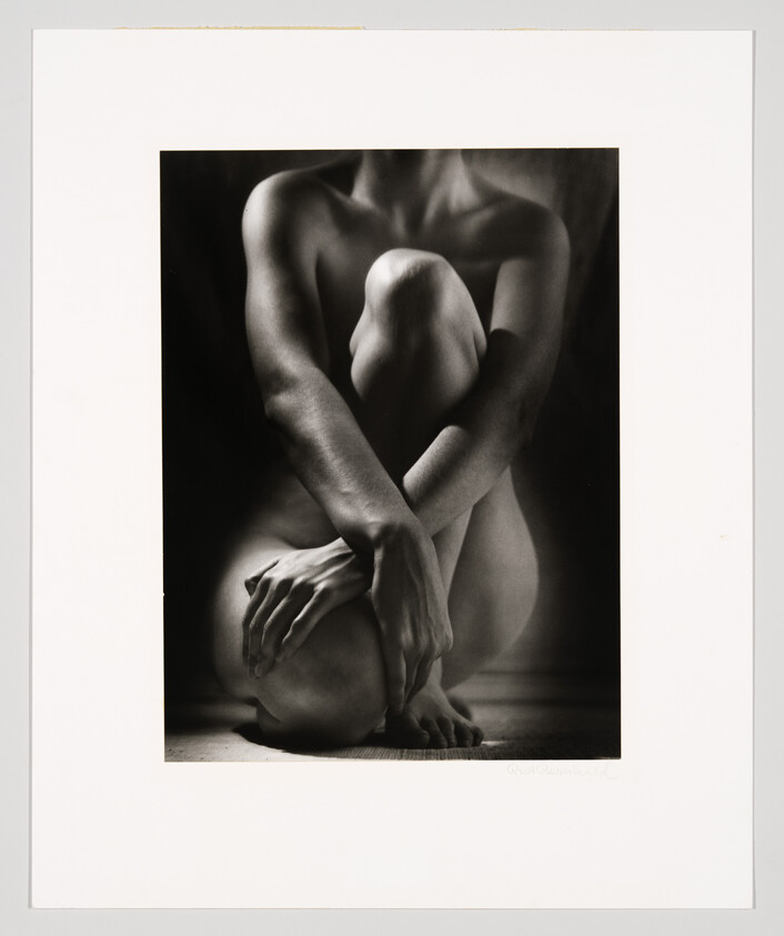 A seated nude person curls their knees to their chest with arms wrapped around legs in dramatic lighting.
