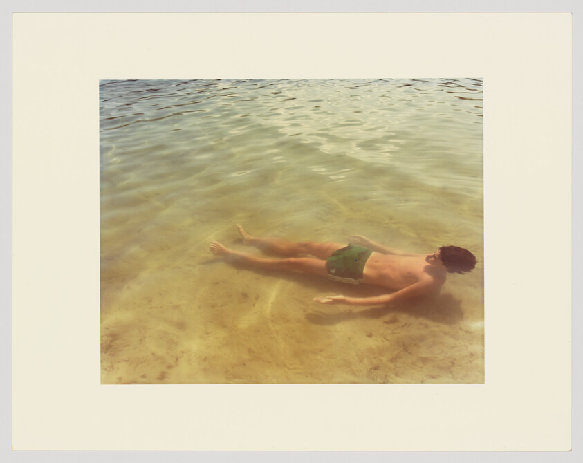 A person floats face-up in shallow clear water wearing green swim trunks.
