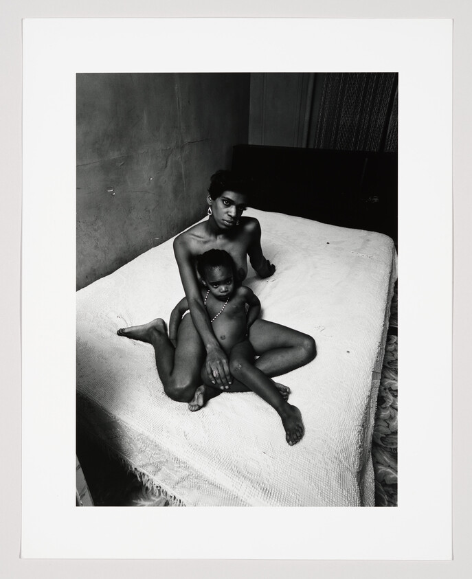 A mother sits on a bed holding her young child closely while looking toward the camera.