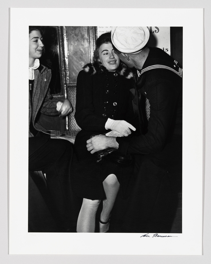 A black and white photograph capturing a tender moment where a sailor in uniform is leaning in to kiss a smiling woman on the cheek, while another woman looks on with a smile. The trio appears to be seated in a subway car, with a sign in the background that reads "Help to keep your subway neat." The image is signed by the photographer at the bottom right.