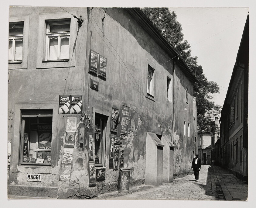 A black and white photograph depicting an old street scene with a man in a suit walking away from the camera. The buildings on the left are adorned with various vintage advertisements and posters, including brands like Persil and Maggi. The architecture appears aged and the street is narrow, lined with cobblestones and framed by the shadows of a sunny day.