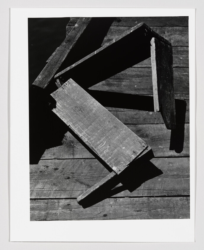 Two broken wooden stools lie on a sunlit wooden deck casting strong diagonal shadows.