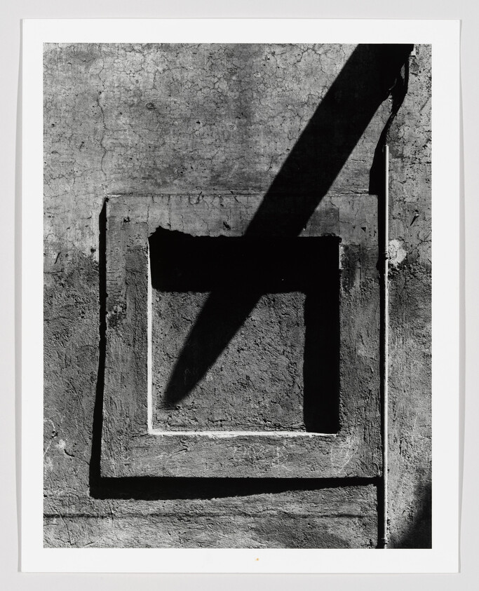 Sun casts a bold diagonal shadow across a recessed square panel on a textured concrete wall.