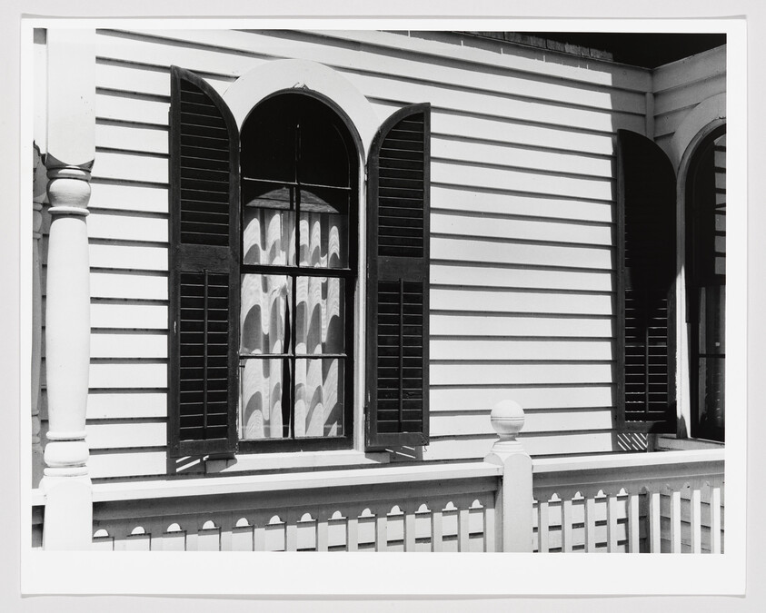 Black and white photo of a window with open shutters on a striped wooden house, with a partial view of a column and railing.