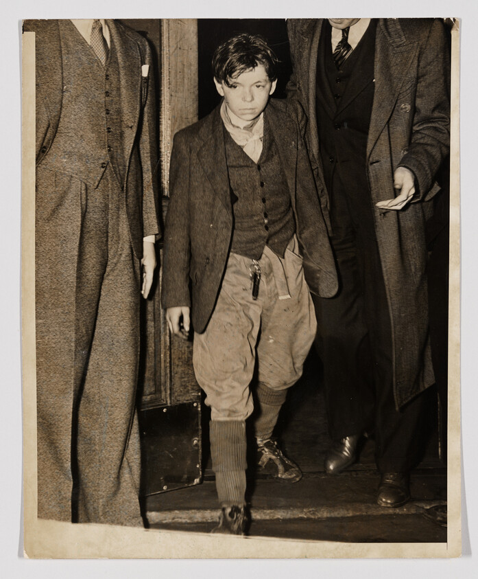 Young boy in worn clothes is being escorted by two men down a doorway.