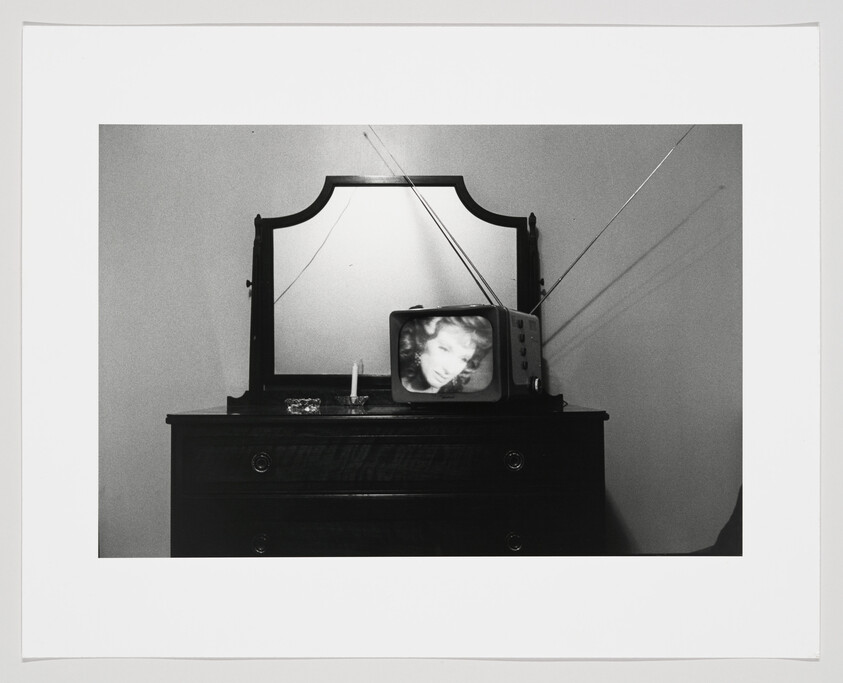 A black and white photograph depicting a vintage television set on top of a dark wooden dresser with a mirror. The TV screen displays the blurred image of a woman's face, and there is a single lit candle and a small box on the dresser. The mirror reflects the TV and the candle, creating a symmetrical effect with light rays emanating from the candle's reflection.