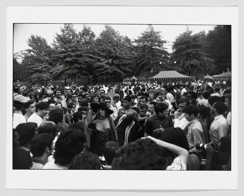 A woman wearing a wide hat and corset stands in a crowded park as people photograph her.