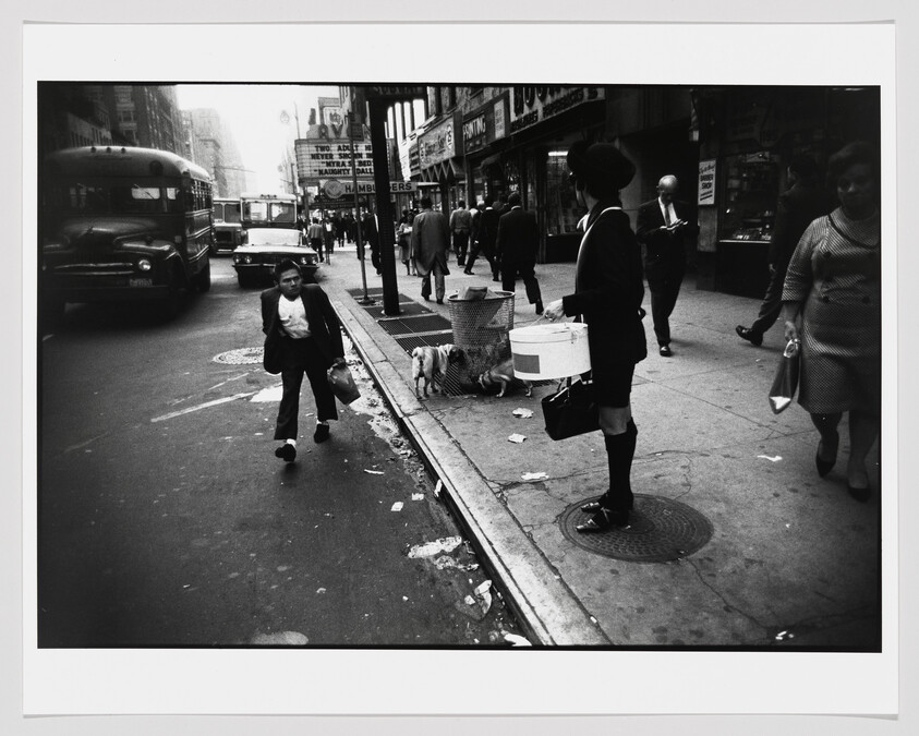 A black and white street photograph capturing a bustling city scene with pedestrians. On the left, a young boy walks towards the camera carrying a bag, with a small dog behind him tied to a street vendor's cart. In the center, a woman stands on the sidewalk wearing a dark outfit with a knee-length skirt, high socks, and a hat, looking off to the side. Various other people are seen in the background walking or standing, and there are vintage buses and cars on the street to the left. The image has a dynamic, candid quality typical of mid-20th-century street photography.