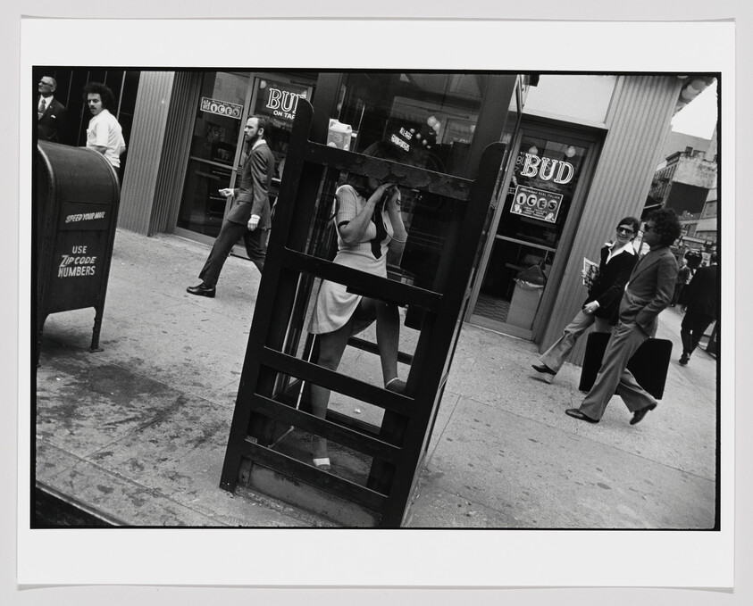 A woman stands inside a street phone booth using a payphone as people pass by.