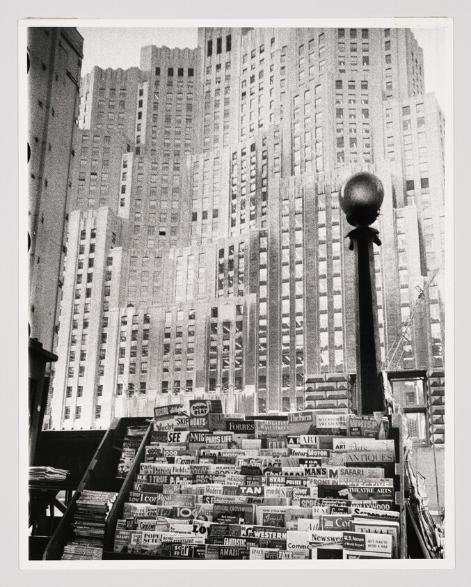 A crowded newsstand filled with magazines stands in front of tall Art Deco skyscrapers.