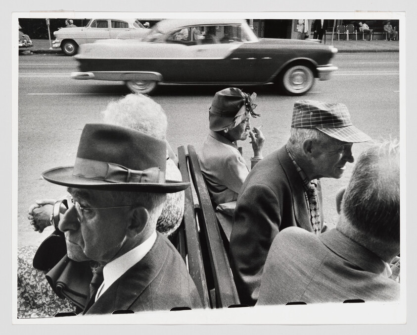 A group of elderly people sit on a bench by a busy street as a car speeds past.