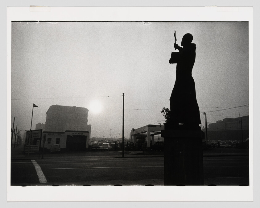 Dark silhouette of a statue holding a cross-like object overlooking a foggy street and buildings.