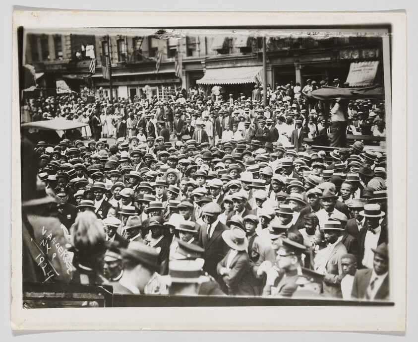A large crowd of men and women wearing hats fills a city street during an outdoor gathering.