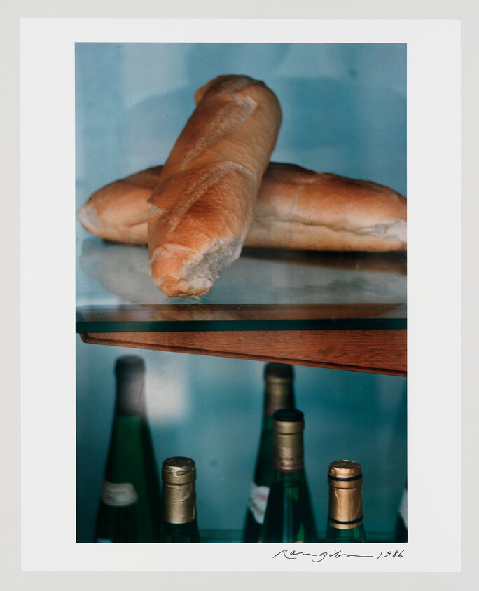 Two baguettes rest on a glass shelf above several wine bottles.