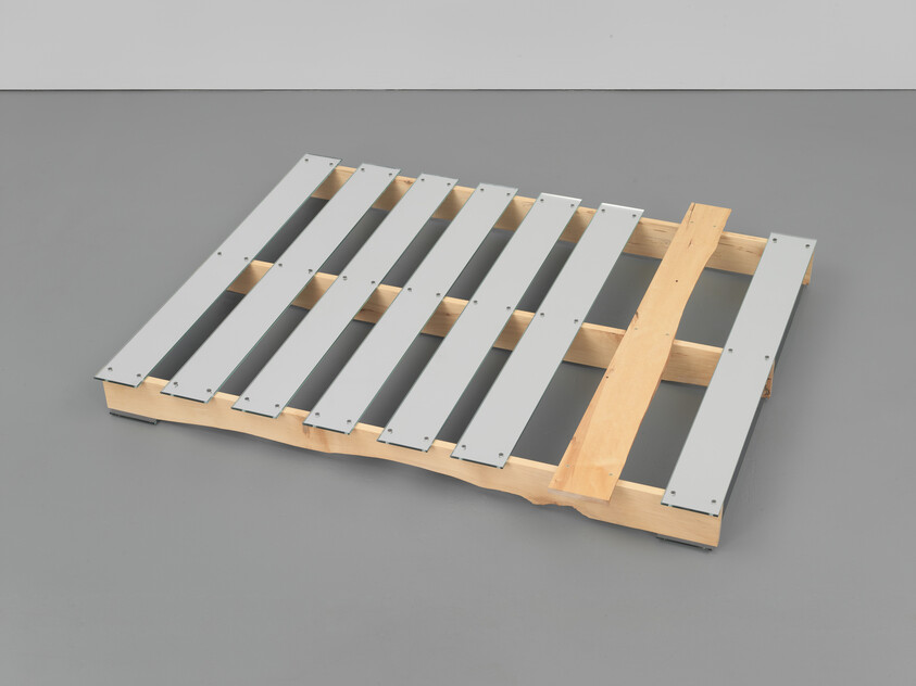 Wooden shipping pallet with evenly spaced metal slats resting on a gray floor.