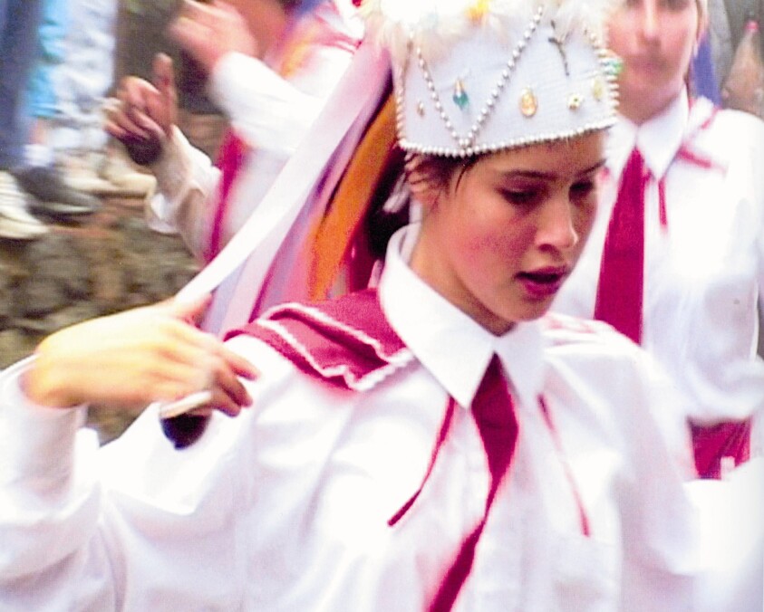 Young woman wearing a decorative crown and white shirt with red tie dances in a procession.