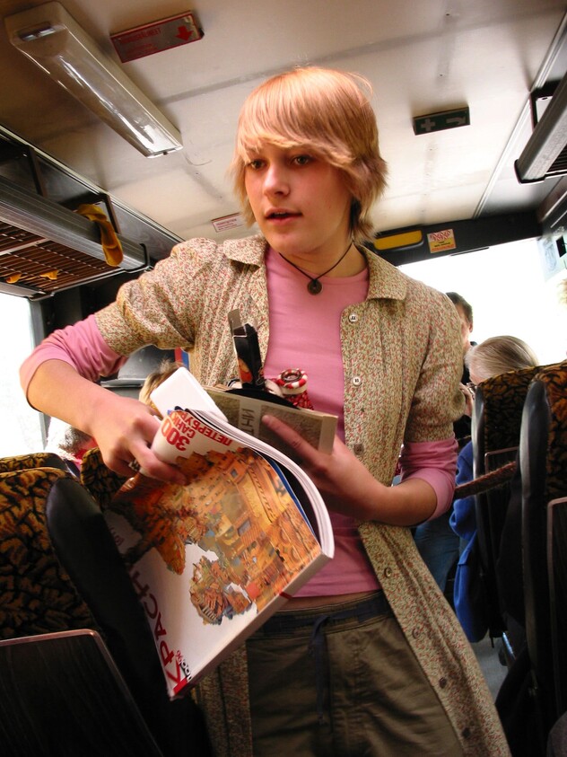 Teen standing on a bus holding open travel guides and magazines while talking.