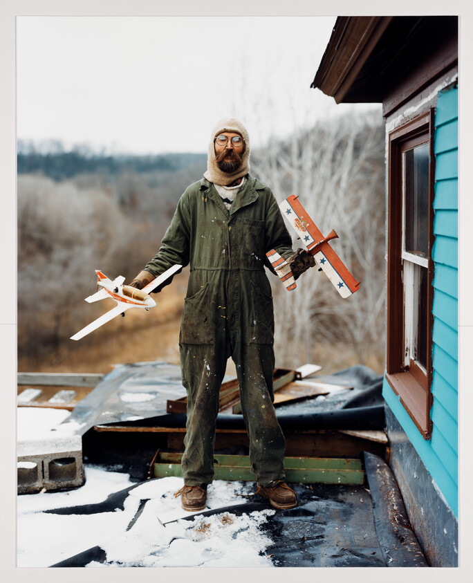 Bearded man in green coveralls stands on a snowy roof holding two toy balsa airplanes.