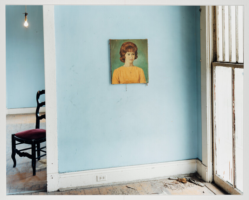 Portrait of a woman hangs on a pale blue wall in a sparsely furnished, dusty room.