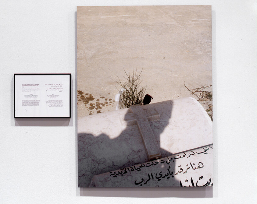 Framed artwork showing a sunlit tombstone with a cross and Arabic inscription on a white wall.