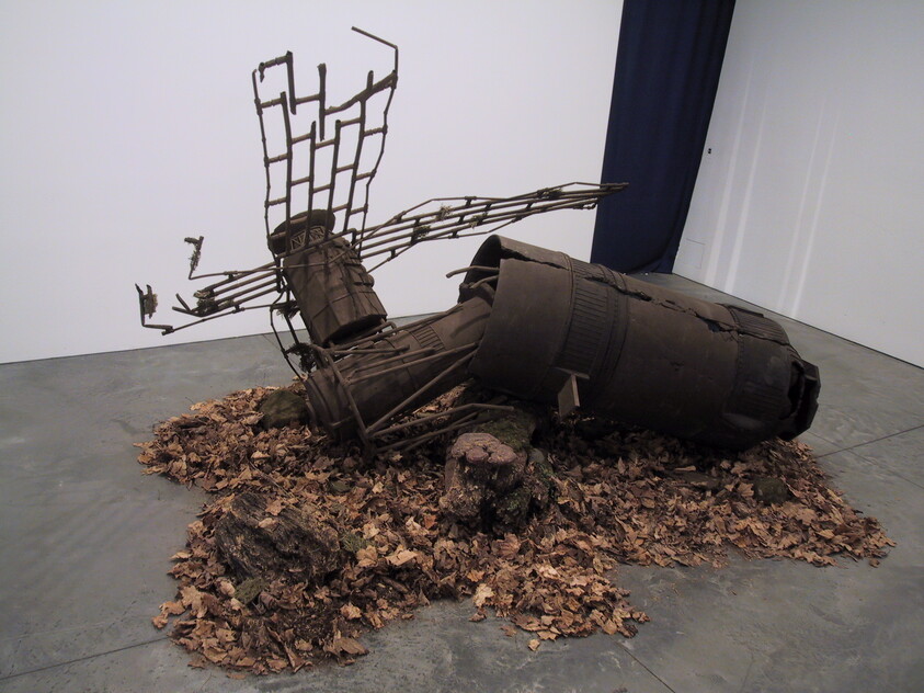 A sculpture resembling a fallen tree made from metal and other materials lies on a bed of dried leaves and branches, with its roots and branches splayed out, inside an art gallery with grey flooring and white walls.