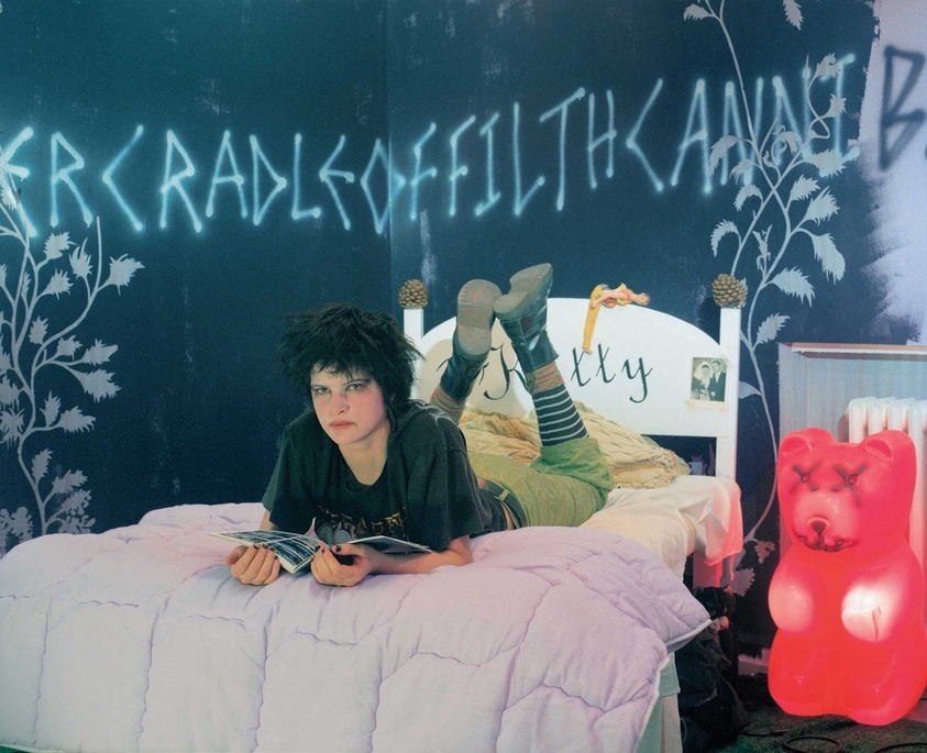 Person with short hair lies on a bed, reading. Neon text on the wall, striped socks, and a red bear-shaped lamp add to the room's decor.