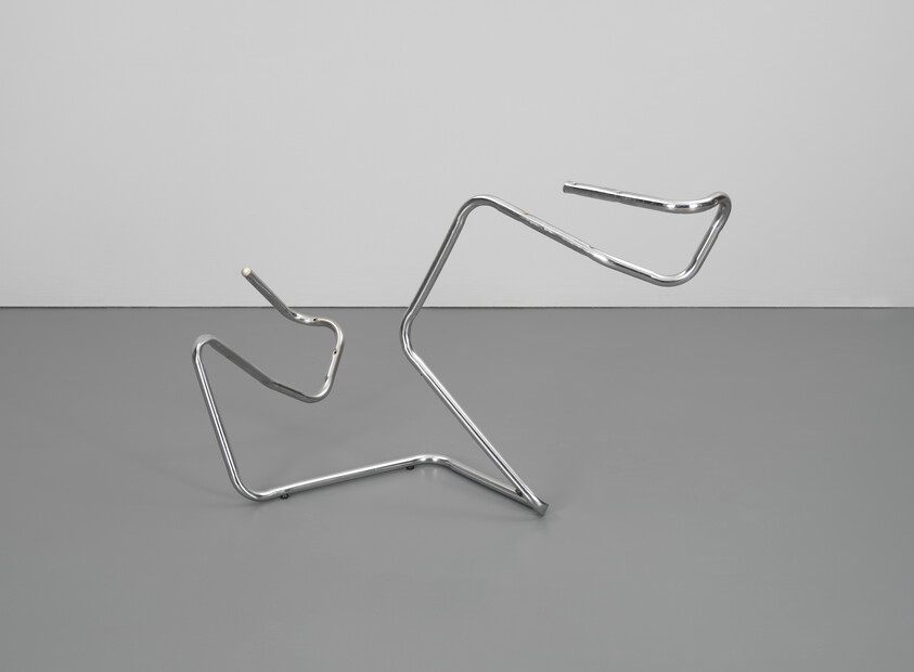A chrome bent metal sculpture forms angular zigzag loops resting on a gray floor.