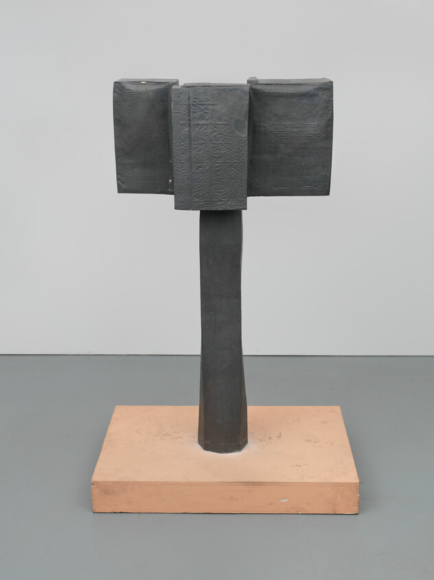 A dark geometric T-shaped sculpture standing on a square wooden base in a gallery.