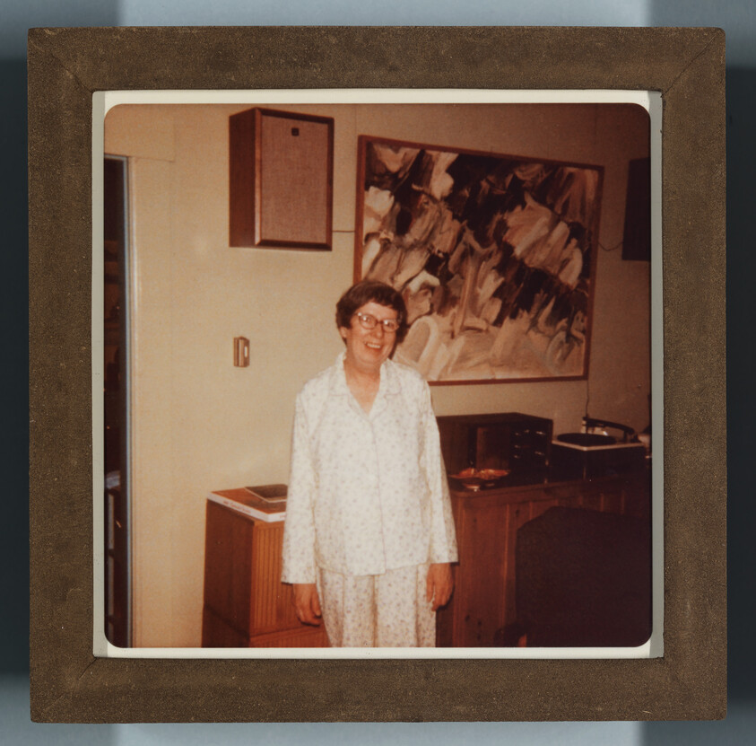 Elderly woman in pajamas smiling while standing in a living room beside a sideboard and painting.