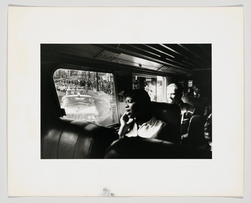 A woman sits on a bus looking thoughtfully out the window as cars and a crowd pass by.