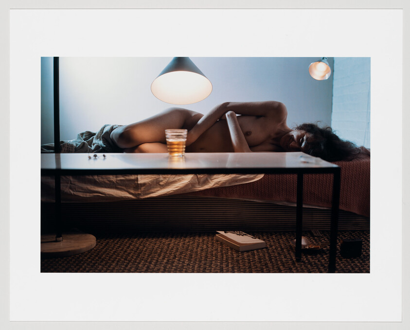 A person lies curled on a bed beside a low table with a glass and hanging lamps.