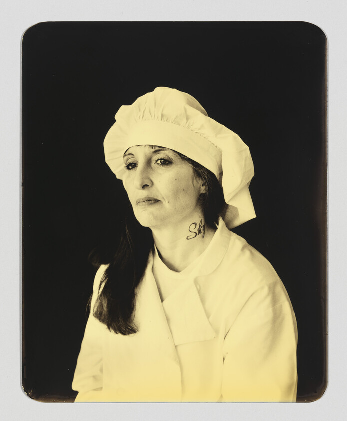 A woman in a chef's hat and coat looking thoughtful, with a small neck tattoo.