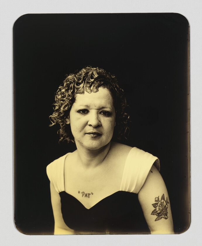 Woman with curly hair wearing a sleeveless dress showing a "PAT" chest tattoo and floral arm tattoo.