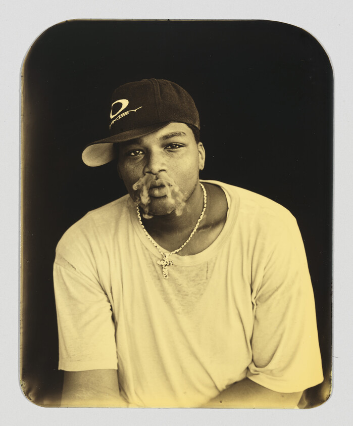 A young man wearing a baseball cap and cross necklace blows smoke from his mouth.