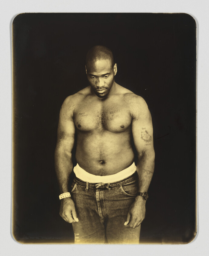 A shirtless man stands looking down with his hands at his sides, wearing jeans and wristbands.