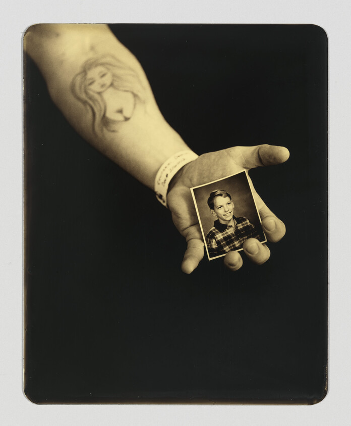 A tattooed hand wearing a wristband holds a small black-and-white school portrait of a smiling boy.