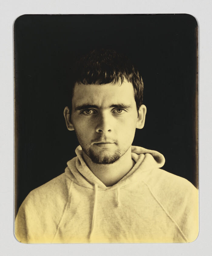 A young man in a hoodie stares directly ahead with a neutral expression in sepia tones.