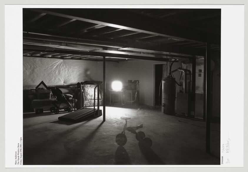Dim basement with a tricycle in the foreground, a treadmill to the left, and a bright lamp on a table.