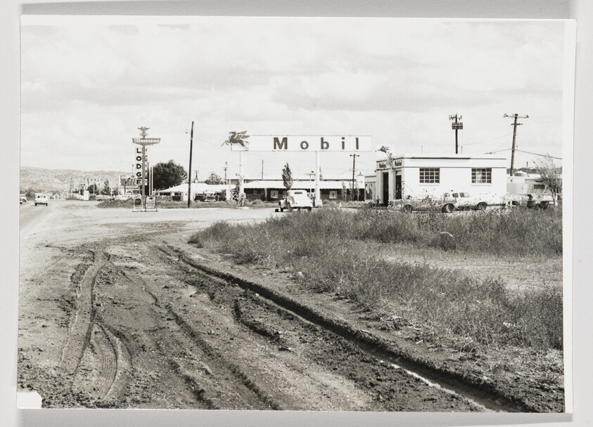 A muddy dirt road leading toward a Mobil gas station and small roadside buildings.