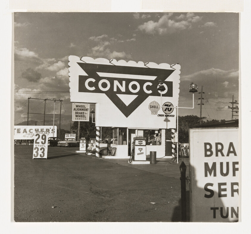 Large Conoco gas station sign over vintage service pumps with fuel price signs visible.
