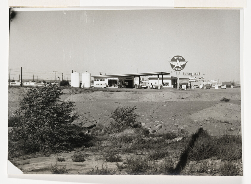 A Flying A Service gas station with canopy and pumps sits beyond a dry scrubby field.