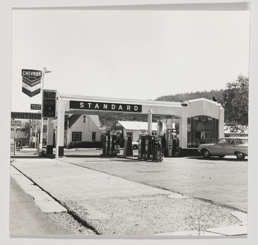 Vintage Standard gas station with fuel pumps under the canopy and a parked car by the service bay.